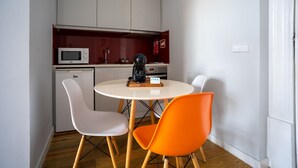 Eine kompakte Küche mit Mikrowelle, Kaffeemaschine und einem kleinen Kühlschrank. Es gibt einen runden Tisch mit zwei Stühlen, einer weiß und einer orange.