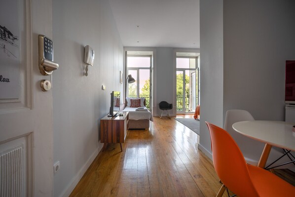 Ein modernes Zimmer mit Holzboden, einem Bett, einem Schreibtisch und einem Esstisch mit orangefarbenen Stühlen.