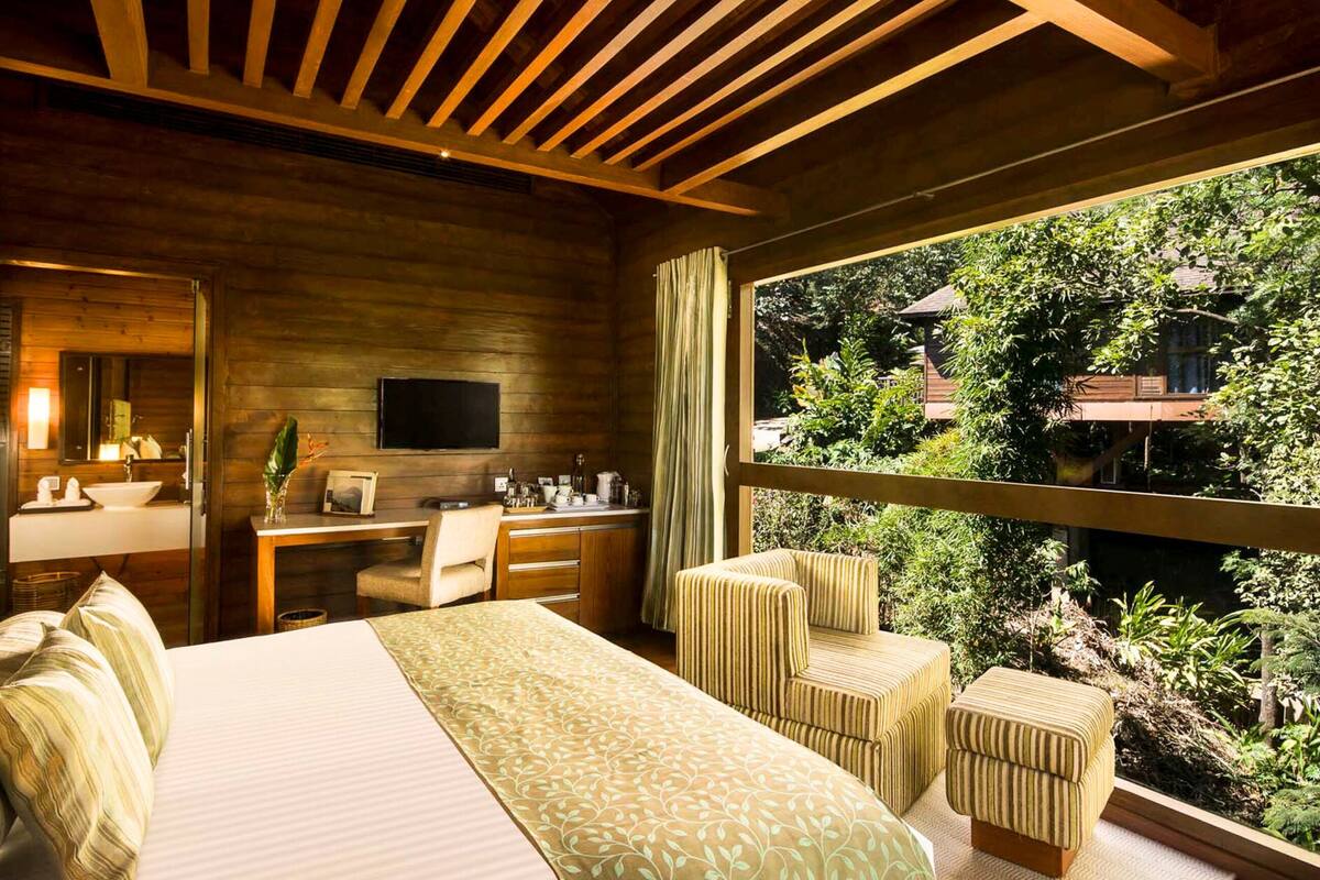 A bedroom with a large bed, a desk, and a chair, a TV, and a window with a view of greenery and a building.