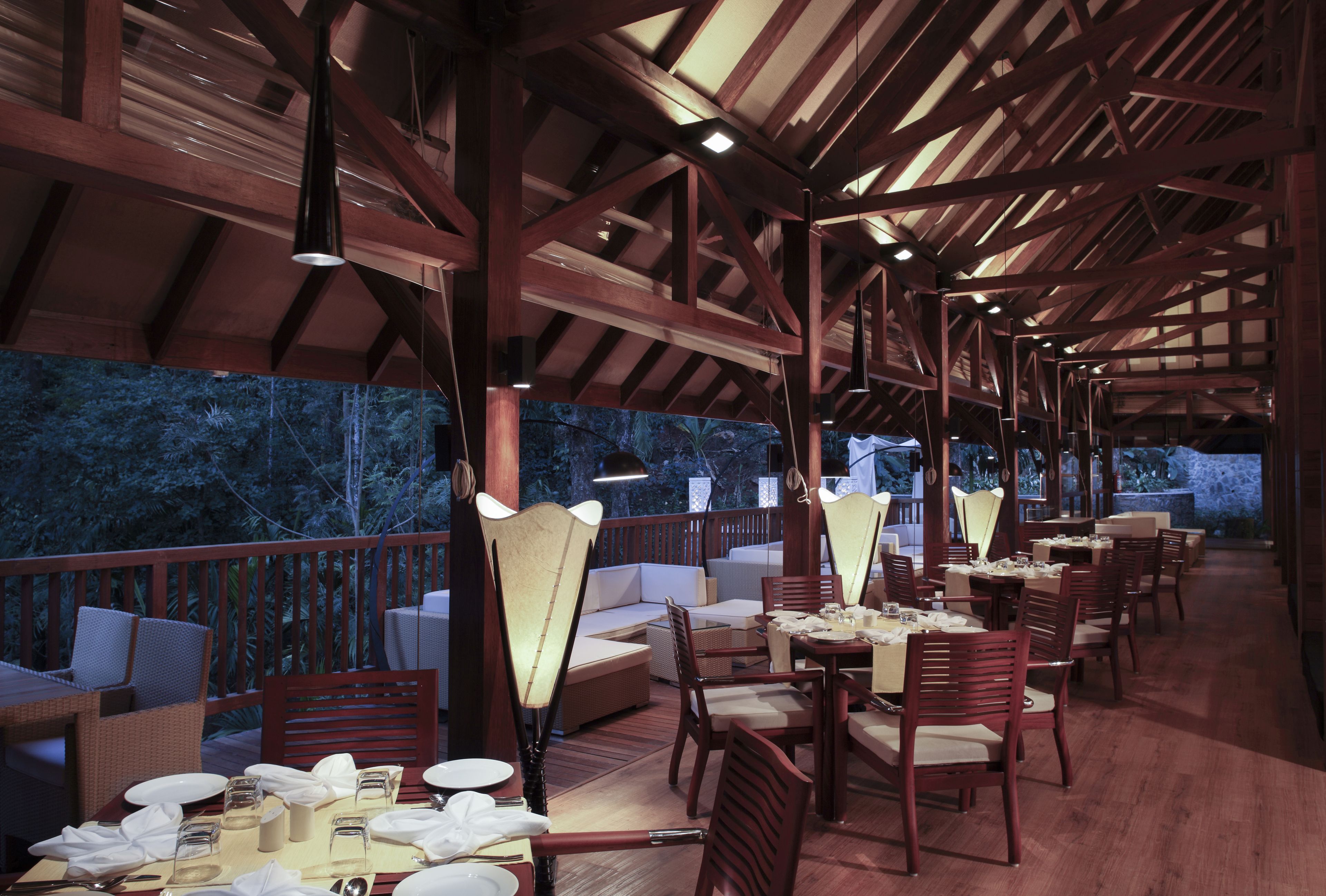 Un área de comedor al aire libre con vigas de madera, mesas, sillas y lámparas colgantes.