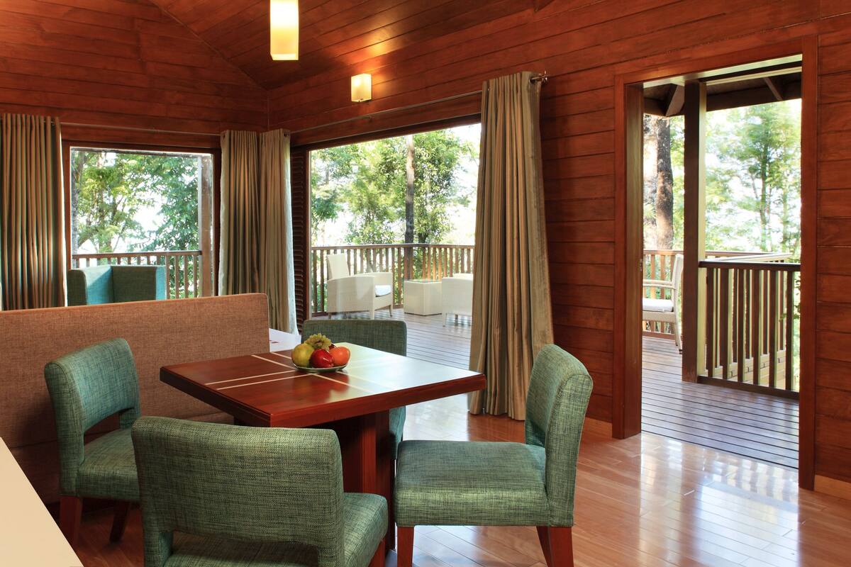A wooden cabin with a dining area featuring a table and chairs, a kitchen area with a kettle and mugs, and large windows with curtains.