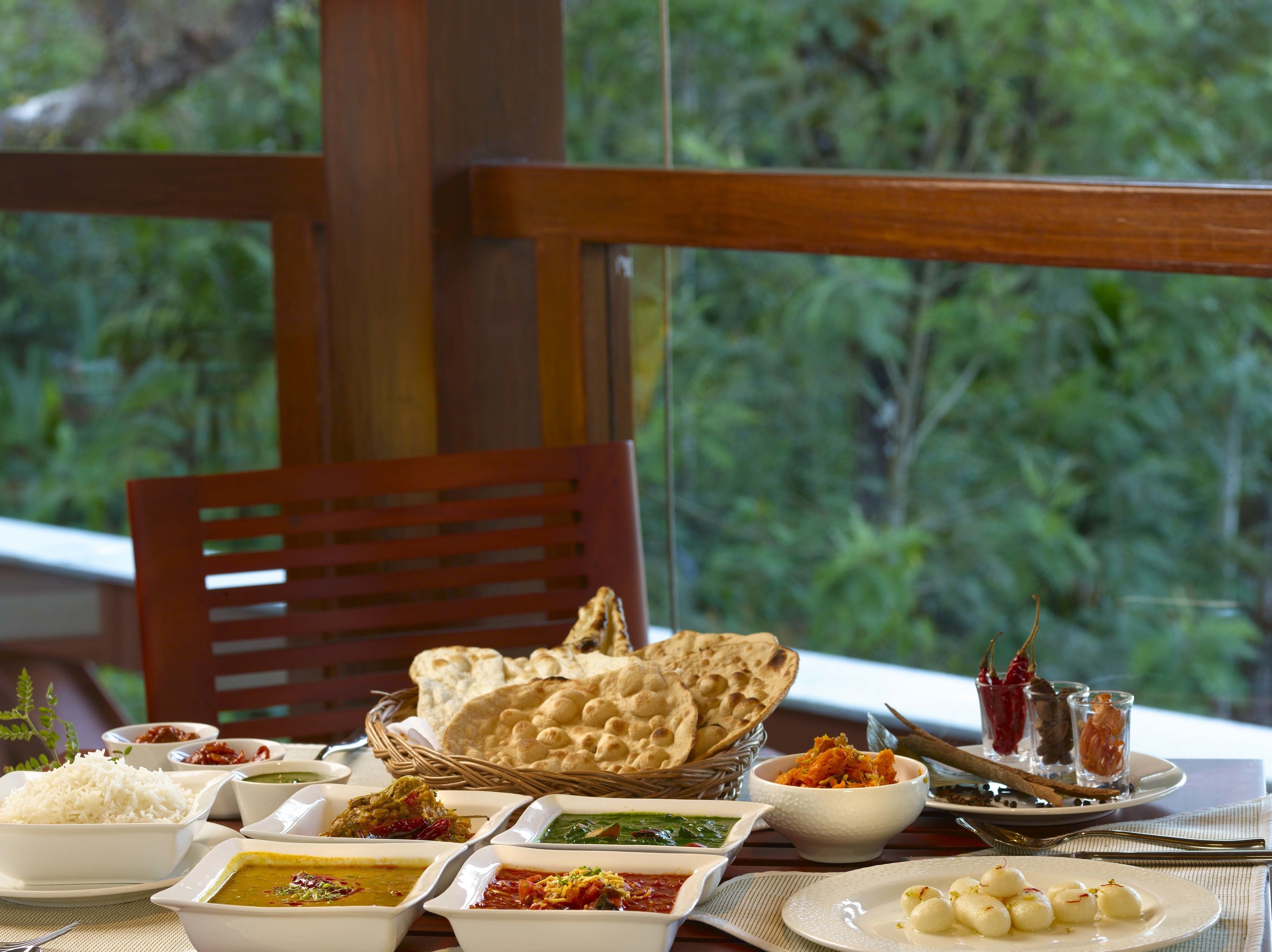 A table set with various Indian dishes, including rice, curry, and bread, with a view of greenery in the background.