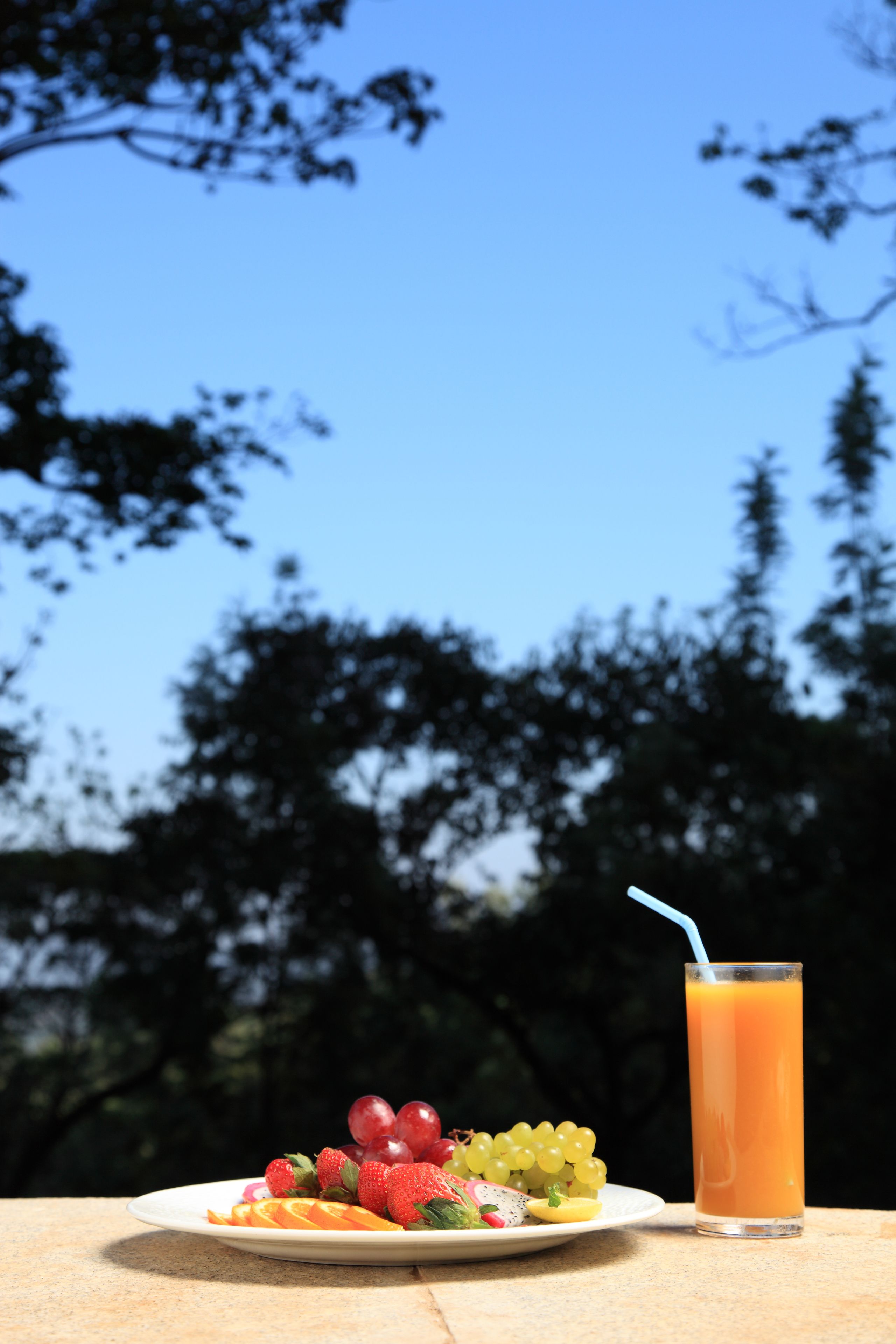 Un plato de fruta y un vaso de jugo de naranja sobre una mesa al aire libre, con un cielo azul despejado y árboles al fondo.