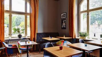 A cozy cafe with wooden tables and chairs, large windows with curtains, and potted plants.