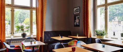 A cozy cafe with wooden tables and chairs, large windows with curtains, and potted plants.
