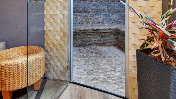 A modern bathroom with a glass shower enclosure, a wooden bench, and a potted plant. - Hotel DaVinci (Marianske Lazne)