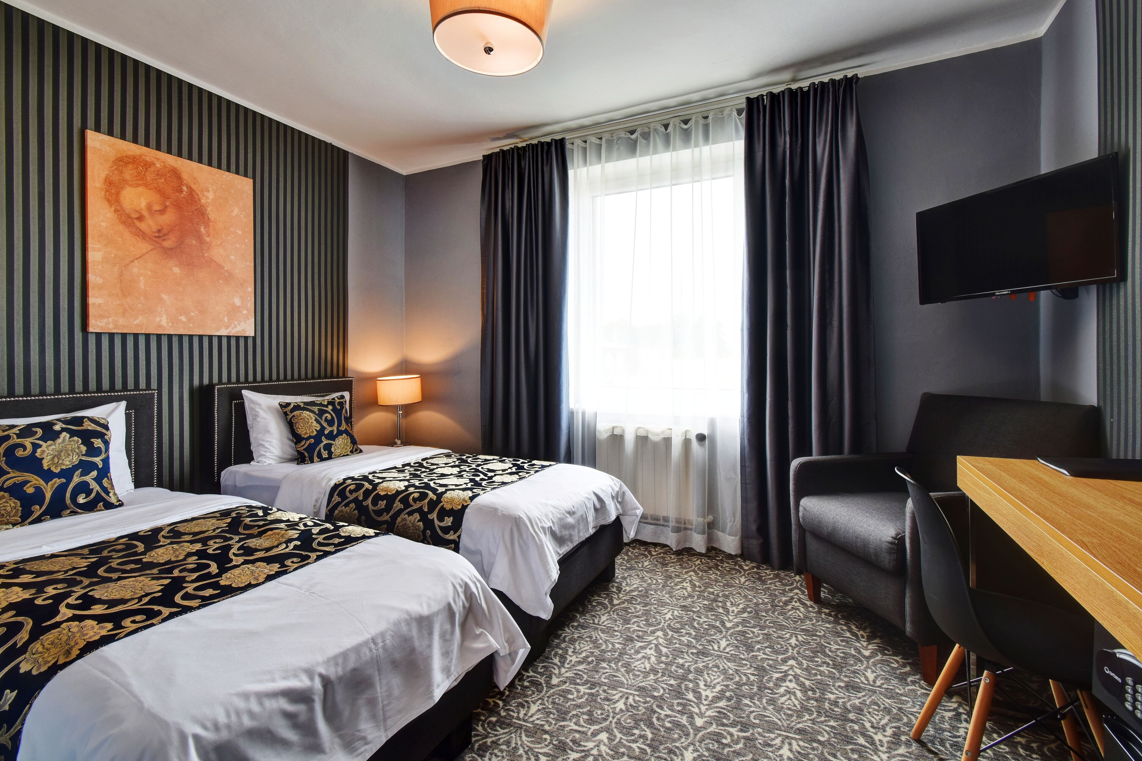 Ett hotellrum med två sängar, ett skrivbord, en stol, en platt-TV och ett stort fönster med gardiner.