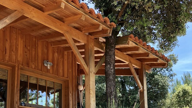 A wooden cabin with a covered porch, a dining area with chairs, and a large tree in the background. - WOODEN HOUSE IN THE BORDER OF THE OCEAN (Soulac-Sur-Mer)