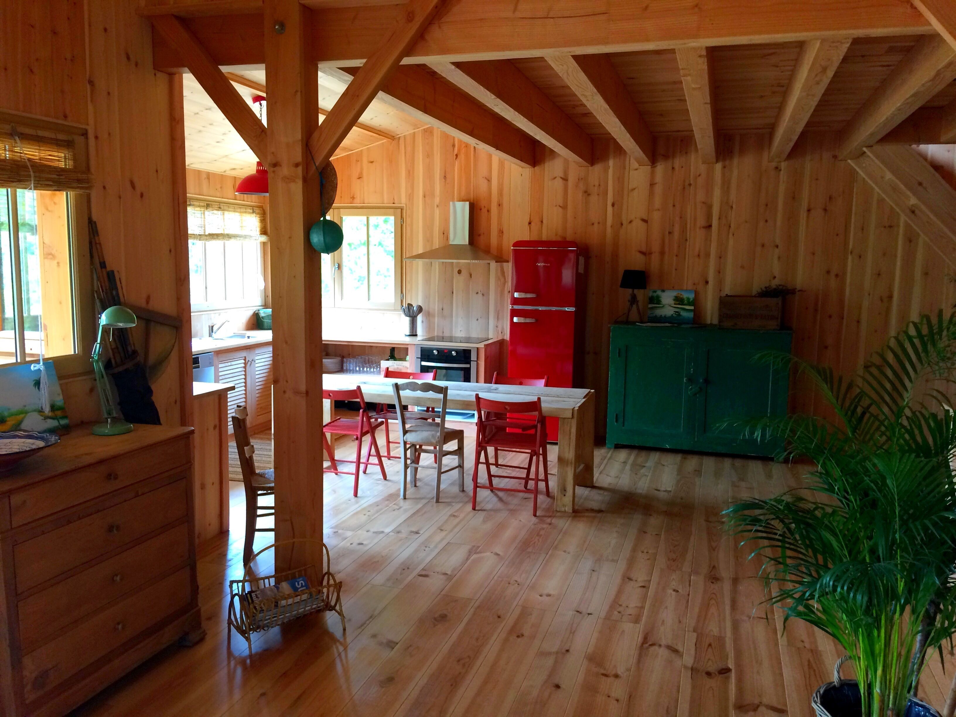 Een houten interieur met een keukenhoek, inclusief een rode koelkast, een eettafel met stoelen en een groen kastje.