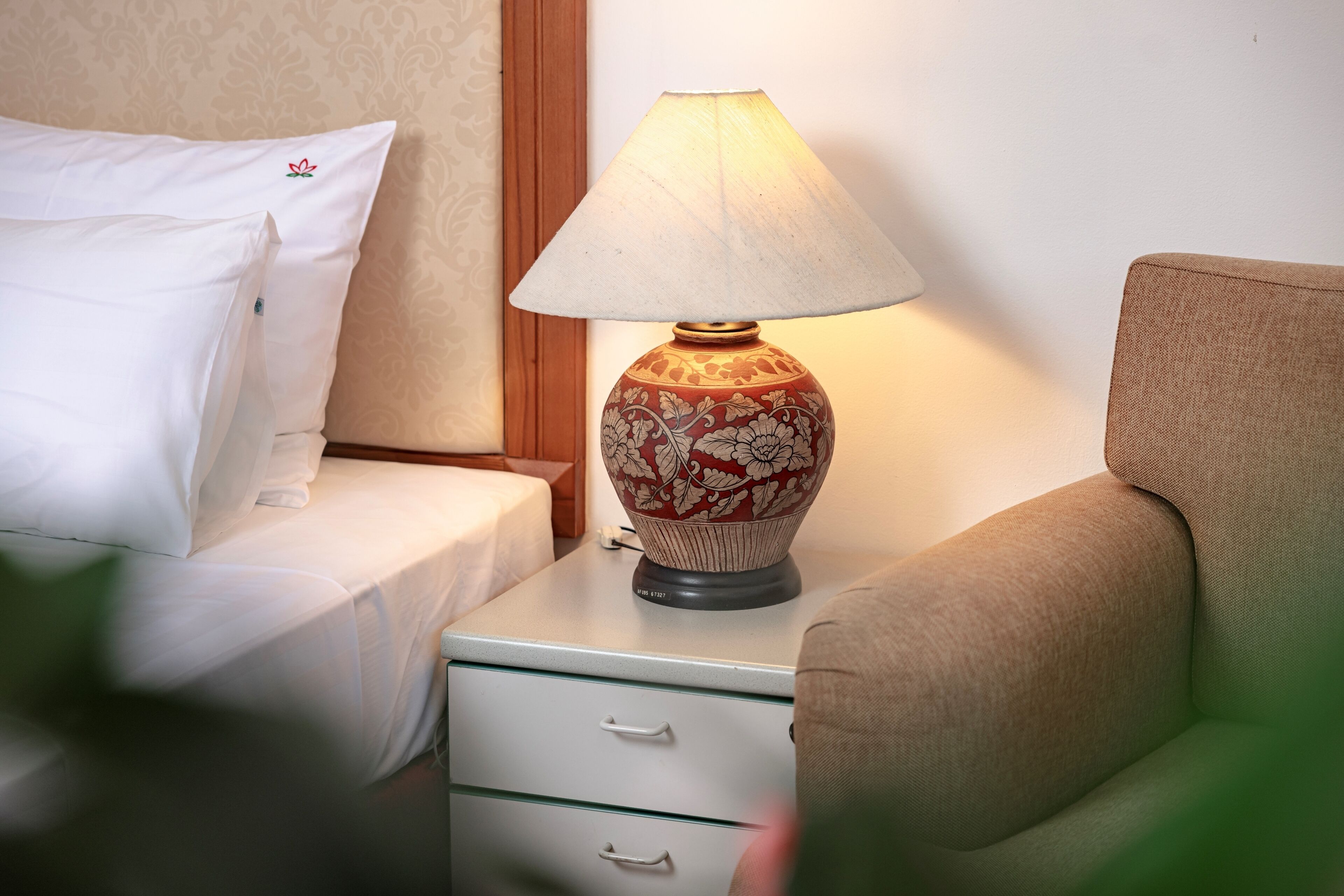 A bed with white bedding and a pillow with a red flower emblem, a bedside table with a floral lamp, and a beige armchair.
