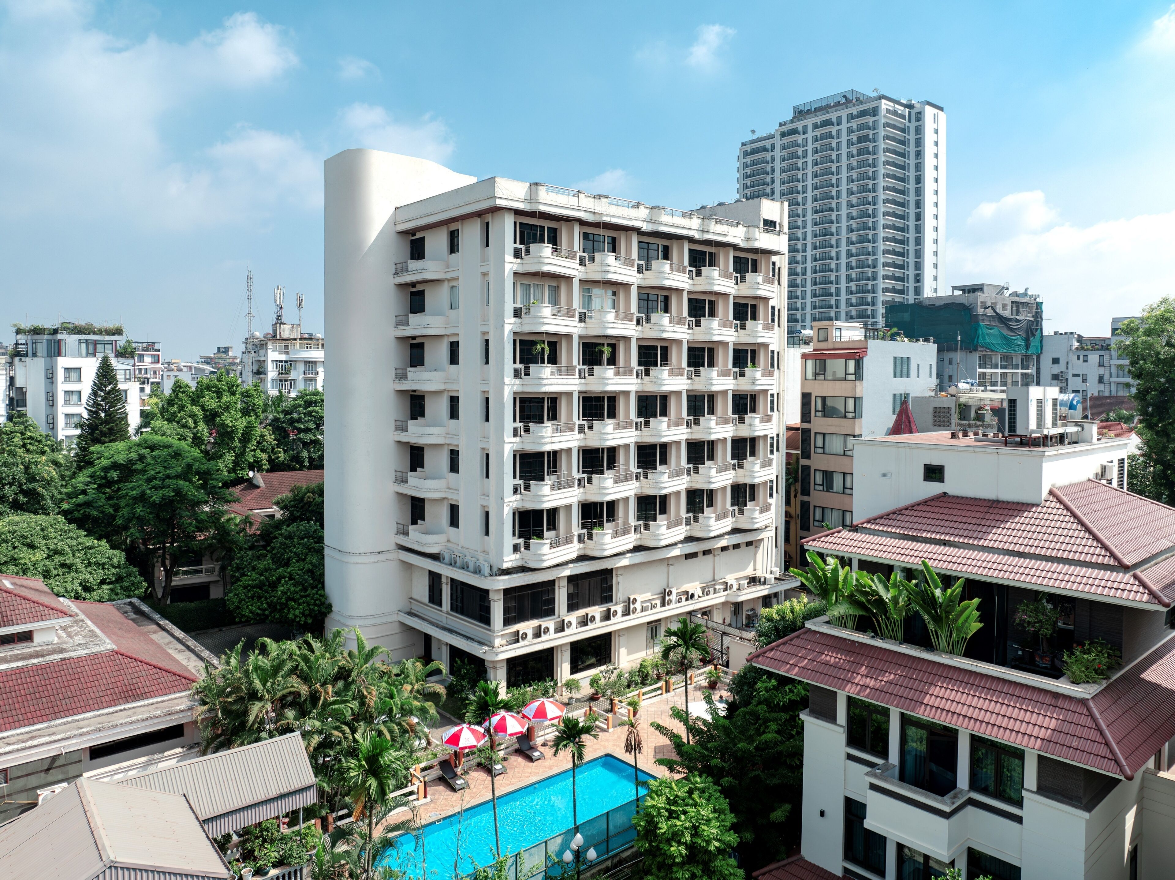 A multi-story hotel with a rooftop pool area, surrounded by other buildings and greenery.