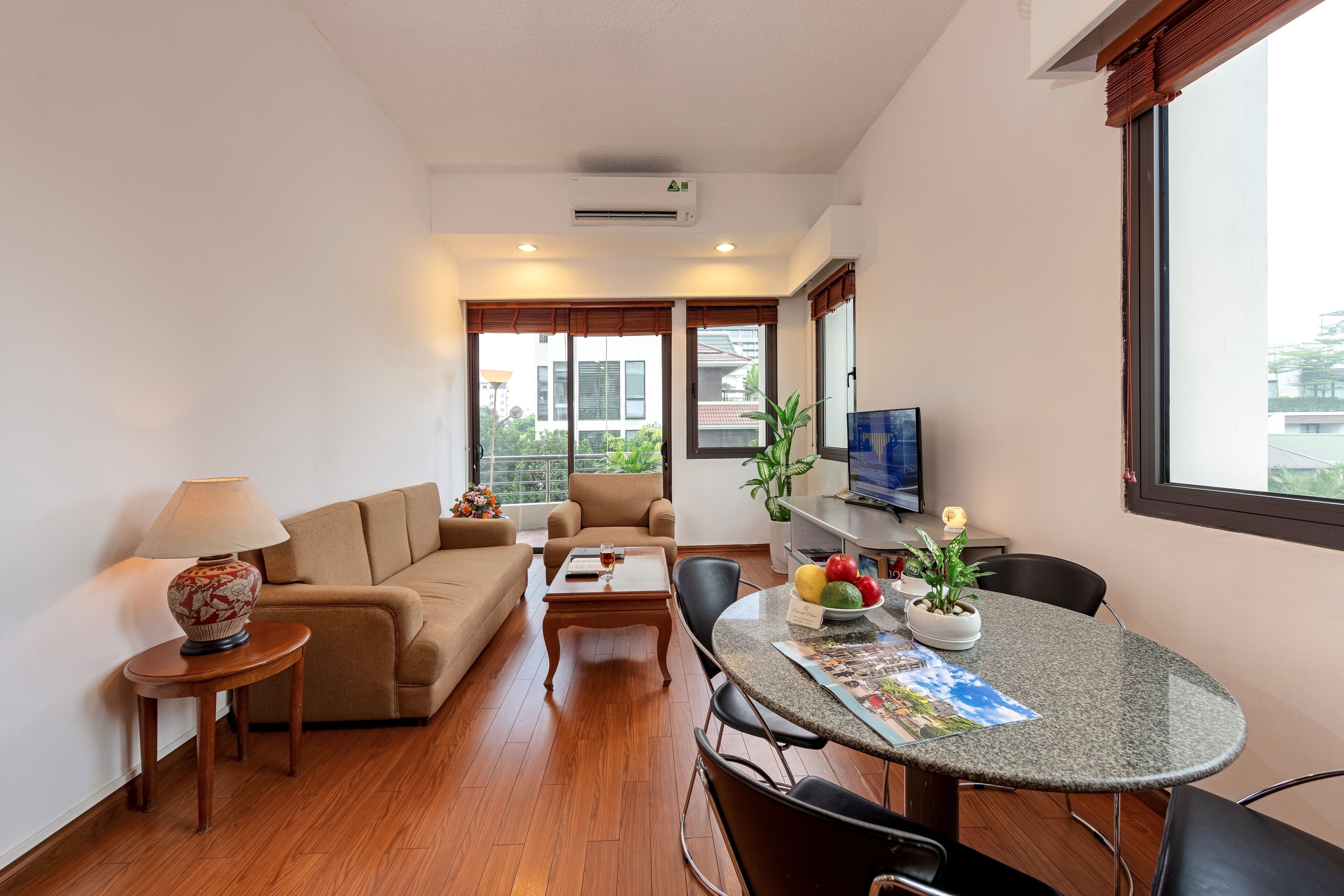 Uma sala de estar moderna com sofá, mesa de centro e mesa de jantar com cadeiras. Há uma televisão sobre um suporte, um abajur e um vaso com planta.
