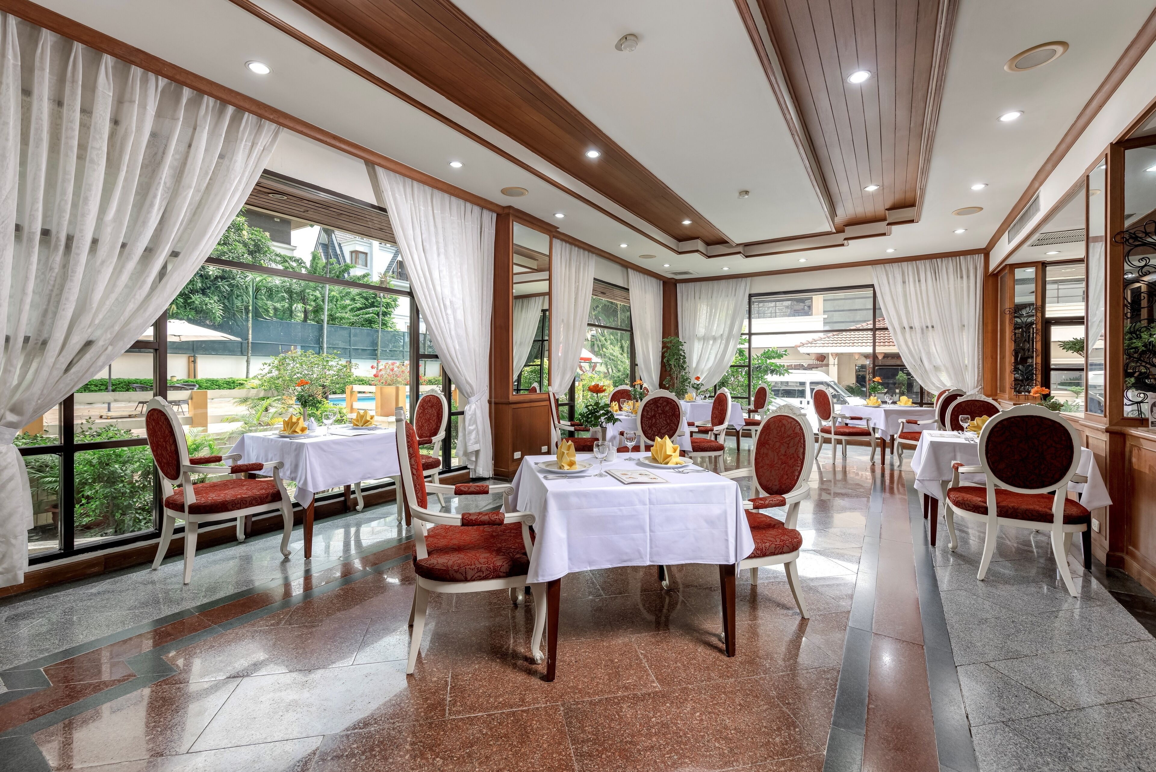 A dining area with tables set for service, red upholstered chairs, and large windows offering a view of the outdoors.