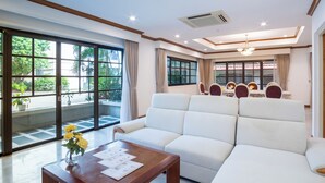 Ampla sala de estar com um sofá branco grande, uma mesa de centro de madeira com um vaso de flores amarelas e uma mesa de jantar posta para seis com cadeiras vermelhas.