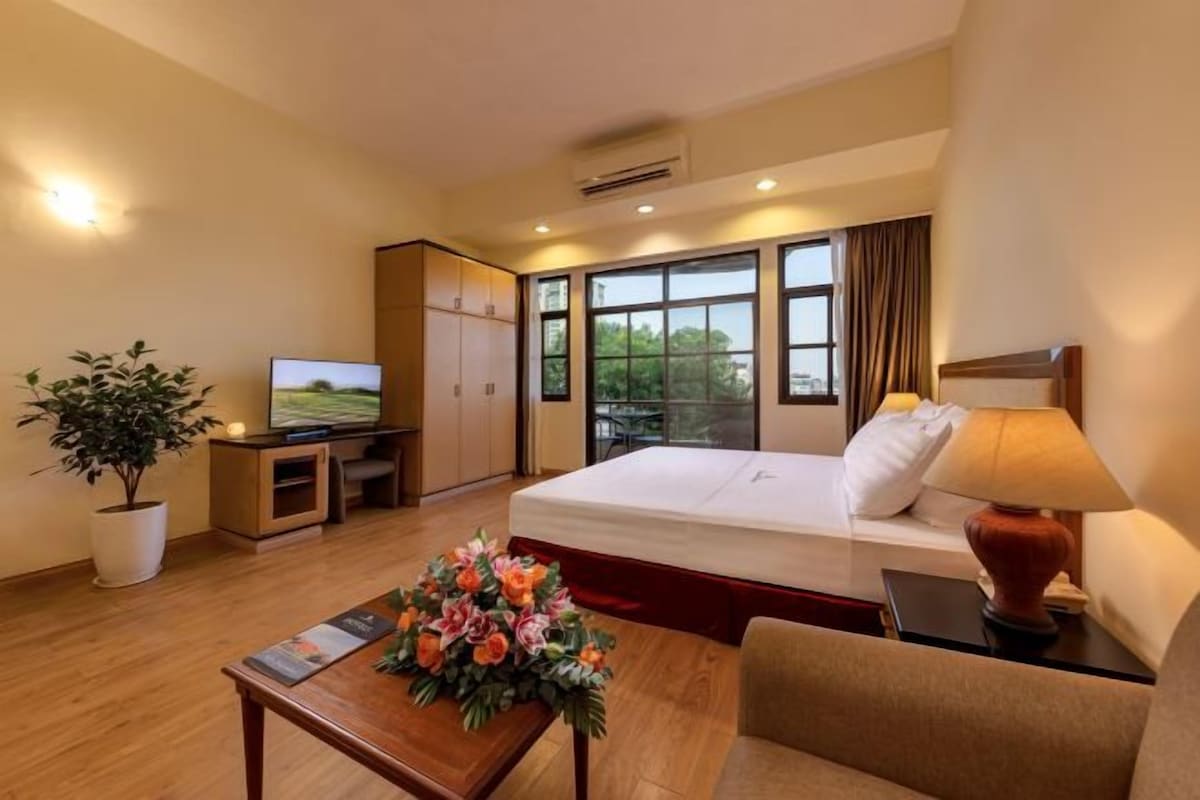 Habitación de hotel con cama, escritorio con computadora, televisor, planta en maceta y ventana con vista a follaje.
