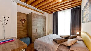 Ein Schlafzimmer mit Holzbalkendecke, einem Bett mit vielen Kissen, einem Nachttisch, einem Kleiderschrank und einem Fenster mit Vorhängen.