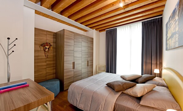 Un dormitorio con techo de madera, una cama con muchos cojines, una mesita de noche, un armario y una ventana con cortinas.