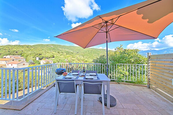 Une table installée à l’extérieur, protégée par un grand parasol, entourée d’une rambarde en métal blanc, et offrant une vue sur un paysage verdoyant.