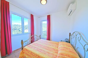 Une chambre à coucher avec un lit, deux fenêtres avec des rideaux, un balcon et un appareil de climatisation.
