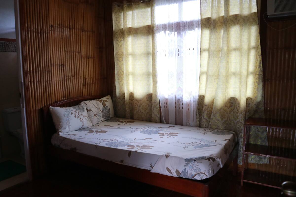 A single bed with floral bedding in a room with bamboo paneling and a window with patterned curtains.