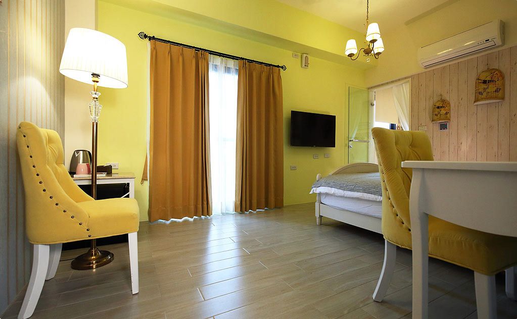 Una habitación con una cama, dos sillones amarillos, una mesa, una lámpara, un televisor y una ventana con cortinas.