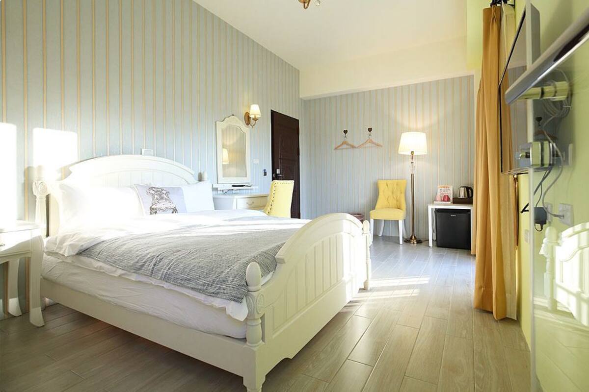 A hotel room with a bed, bedside table, lamp, chair, and a small desk with a kettle.