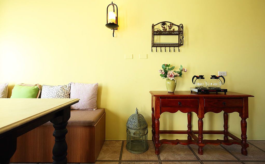 A room with a wooden table, a bench, a small wooden cabinet, a decorative wall hanging, a wall-mounted light fixture, and a small metal birdcage.