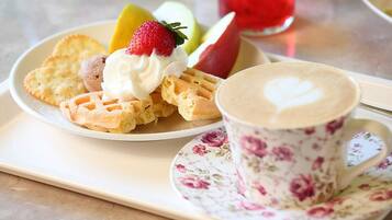 Un plato con waffles, crema batida y fresas, junto a una taza de café con diseño floral.