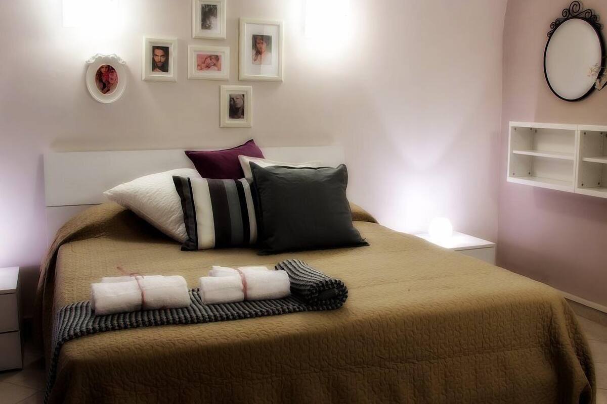 A neatly made bed with a brown bedspread, pillows, and rolled towels. There are framed photos on the wall, a bedside table, and a mirror on the wall.