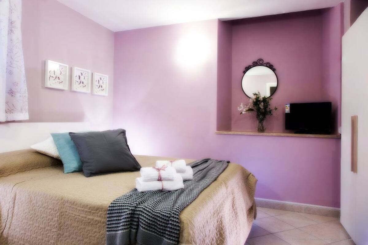 A bedroom with a bed, pillows, a blanket, a mirror, and artwork on the wall.