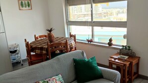 A living room with a grey sofa, a wooden dining table, and chairs. A window with a view of buildings is visible. - T1 - Sea View Balcony (Armação de Pêra)