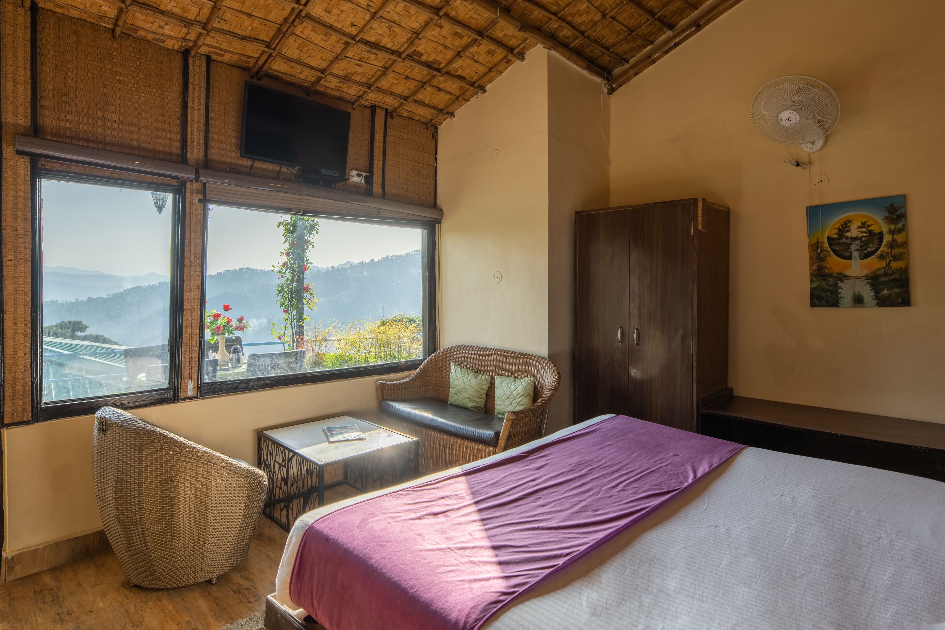 Une chambre à coucher comprenant un lit, une chaise en osier, une petite table, une fenêtre donnant sur les montagnes, une armoire en bois et un ventilateur de plafond.