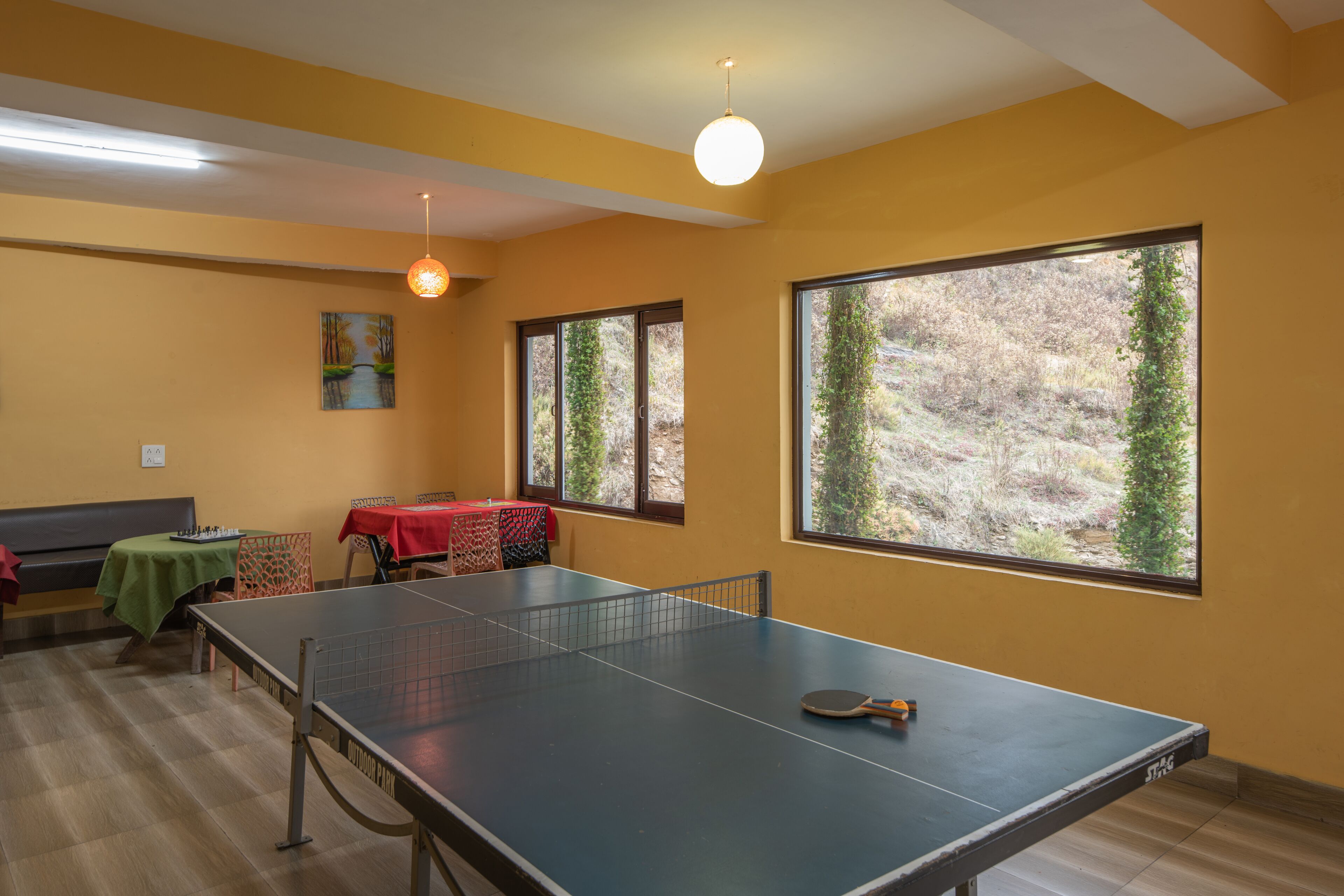 Een kamer met een pingpongtafel, een eettafel met een rode tafelkleed en grote ramen met uitzicht op bomen.