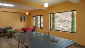 Een kamer met een pingpongtafel, een eettafel met een rode tafelkleed en grote ramen met uitzicht op bomen.