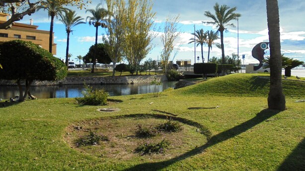A park with a pond, palm trees, and a building in the background. - Los Labradores (Peñiscola)
