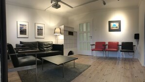 Una sala de estar moderna con un sofá de cuero negro, una mesa de centro de vidrio y un juego de sillones rojo y negro. Hay cuadros enmarcados en la pared, una lámpara y un proyector montado en el techo.