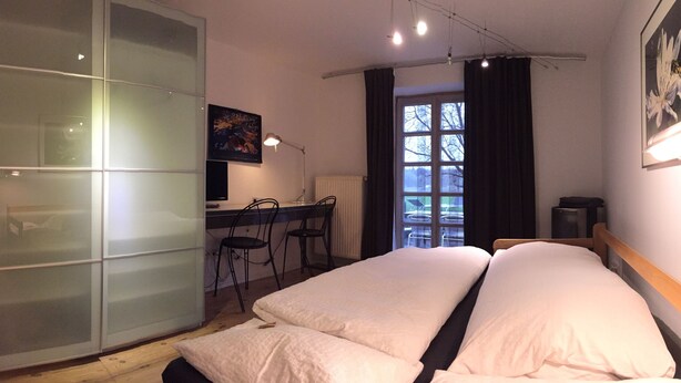 Un dormitorio con cama, escritorio y silla. Hay una ventana con cortinas, un radiador y un reloj en la pared.
