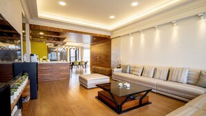 Un moderno vestíbulo de hotel con un mostrador de recepción de madera, un área de descanso con un sofá y reposapiés, y un comedor con sillas y una mesa.