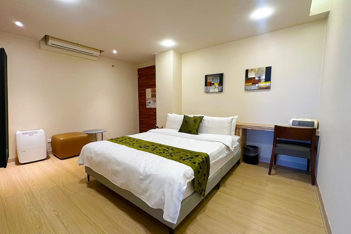 Una habitación de hotel con cama, escritorio, aire acondicionado, cubo de basura y calentador de pared.