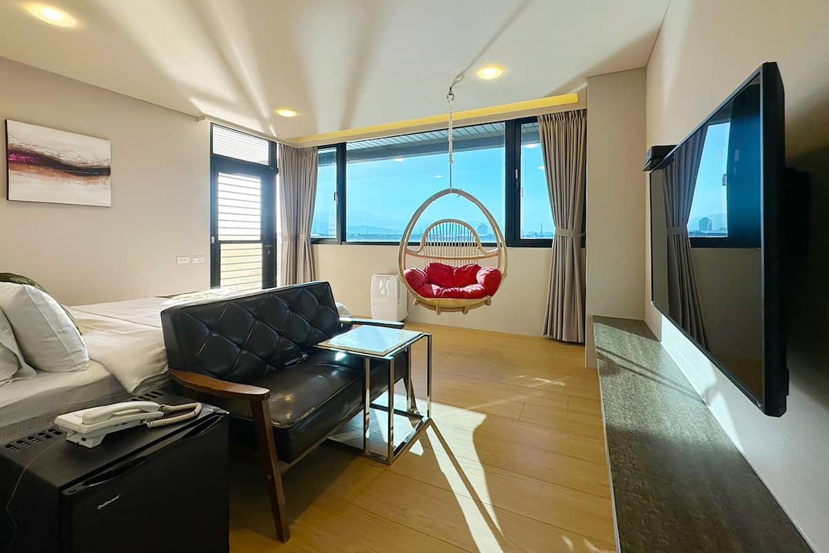 Una habitación de hotel moderna con una cama, un sofá de cuero negro, una silla colgante roja, un televisor de pantalla plana y ventanas amplias con vista a la ciudad.