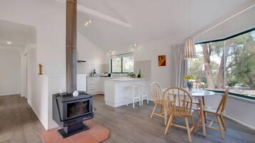 A modern kitchen with a central island, a dining area with wooden chairs, and a large window with a view of trees.
