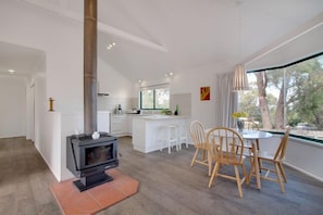 A modern kitchen with a central island, a dining area with wooden chairs, and a large window with a view of trees.