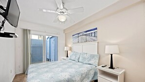 A bedroom with a bed, two bedside tables with lamps, a ceiling fan, a TV mounted on the wall, and a sliding glass door leading to a balcony. - Affordable and ocean front! (San Diego)
