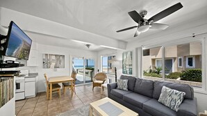 A modern living room with a grey sofa, a wooden coffee table, a dining area with a table and chairs, a ceiling fan, and a TV mounted on the wall. - Affordable and ocean front! (San Diego)