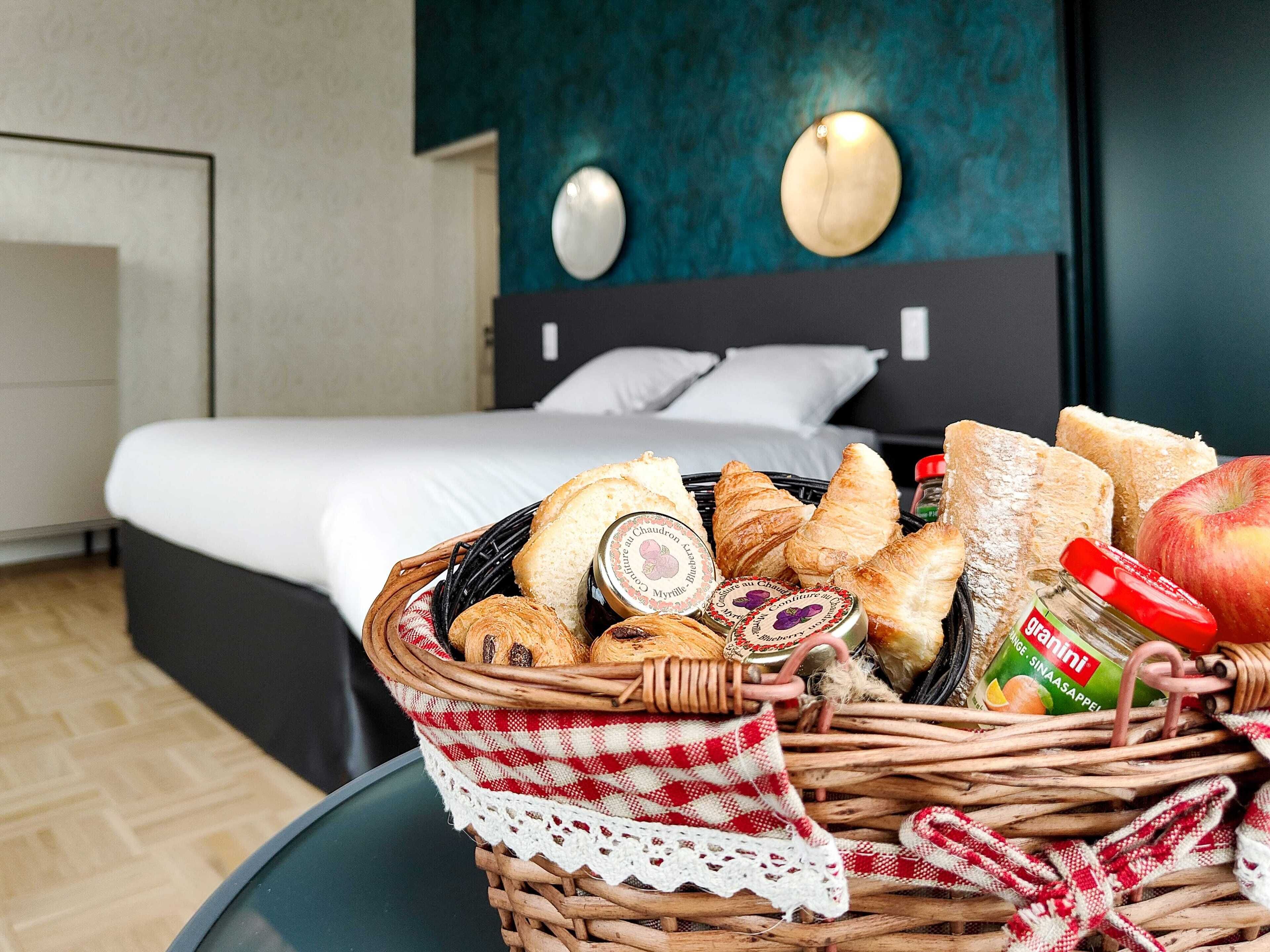 Uma cesta de vime com pão, um pote de geleia e uma maçã sobre uma mesa em quarto de hotel, com uma cama e uma parede escura ao fundo.