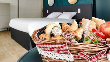 Uma cesta de vime com pão, um pote de geleia e uma maçã sobre uma mesa em quarto de hotel, com uma cama e uma parede escura ao fundo.
