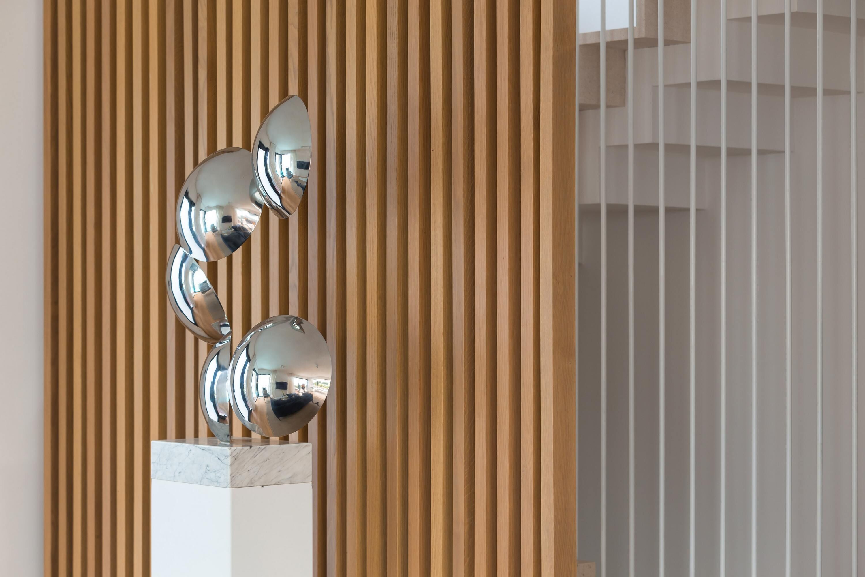 A metallic sculpture with three spherical elements mounted on a marble base, set against a backdrop of vertical wooden slats.