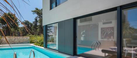 A modern pool area with clear blue water, a lounge chair with a towel, and a stone wall in the background.