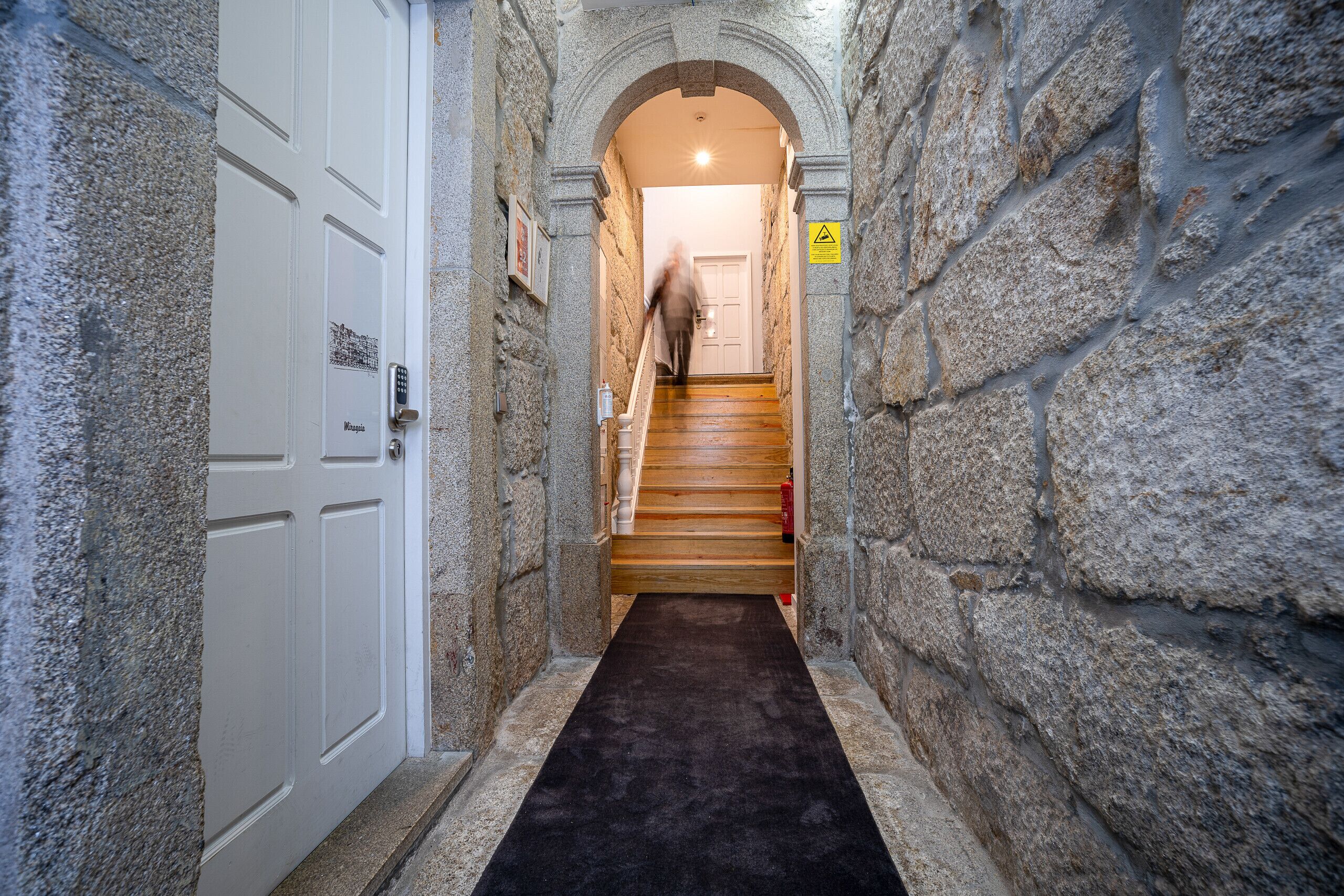 Un étroit couloir en pierre, avec un escalier en bois menant à une porte.