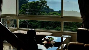 Ein Zimmer mit einem großen Fenster, das einen Blick auf eine Berglandschaft und das Meer freigibt. Auf dem Fensterbrett steht ein Tisch mit einer Obstschale und einem Ventilator.